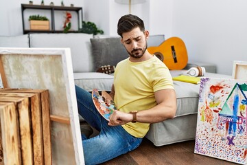 Young man with beard painting canvas at home checking the time on wrist watch, relaxed and confident