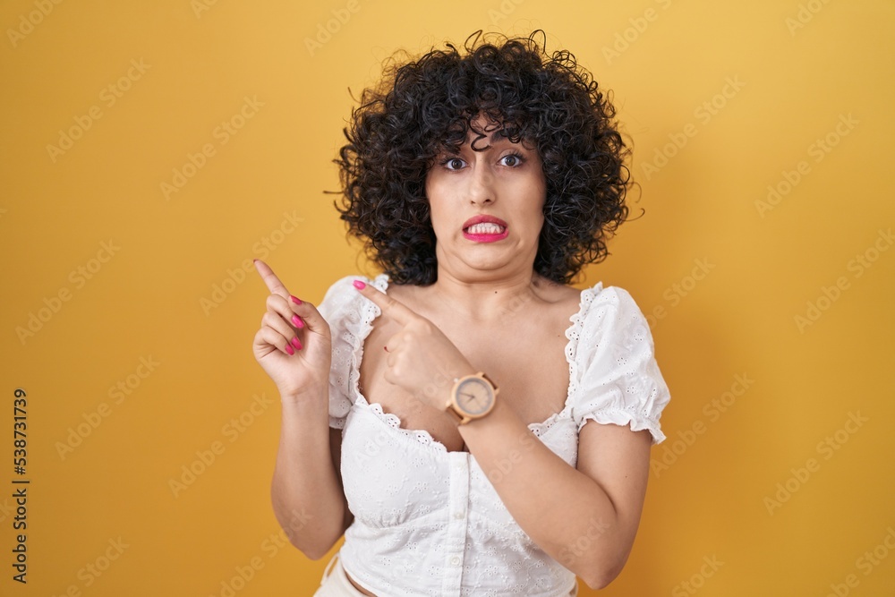 Sticker young brunette woman with curly hair standing over yellow background pointing aside worried and nerv