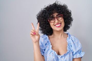 Young brunette woman with curly hair wearing glasses over isolated background smiling with happy...