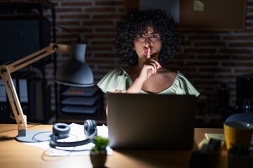 Young brunette woman with curly hair working at the office at night asking to be quiet with finger...