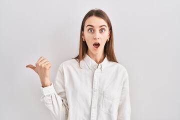 Young caucasian woman standing over isolated background surprised pointing with hand finger to the side, open mouth amazed expression.