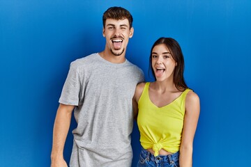 Young hispanic couple standing together over blue background sticking tongue out happy with funny expression. emotion concept.