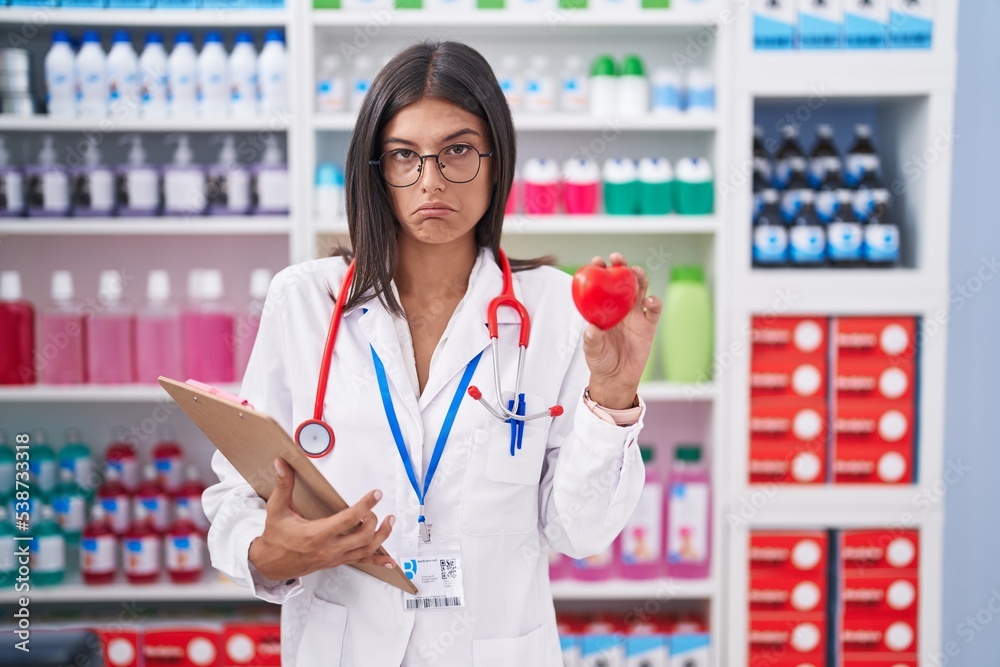 Poster brunette young woman working at pharmacy drugstore holding red heart depressed and worry for distres