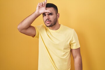 Young hispanic man standing over yellow background making fun of people with fingers on forehead...