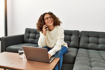 Middle age hispanic woman using laptop talking on the smartphone at home