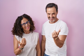 Obraz premium Middle age hispanic couple together over pink background beckoning come here gesture with hand inviting welcoming happy and smiling