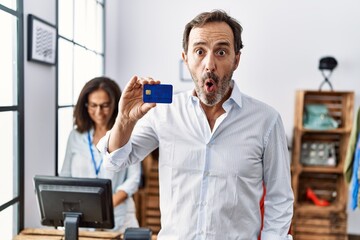 Hispanic man holding credit card at retail shop scared and amazed with open mouth for surprise, disbelief face