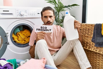 Young hispanic man doing laundry holding eco friendly paper in shock face, looking skeptical and sarcastic, surprised with open mouth