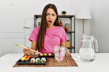 Obraz na płótnie Canvas Young brunette woman eating sushi using chopsticks in shock face, looking skeptical and sarcastic, surprised with open mouth