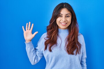 Hispanic young woman standing over blue background showing and pointing up with fingers number five while smiling confident and happy.