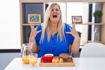 Obraz na płótnie Canvas Caucasian plus size woman eating breakfast at home crazy and mad shouting and yelling with aggressive expression and arms raised. frustration concept.