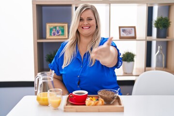 Obraz na płótnie Canvas Caucasian plus size woman eating breakfast at home smiling friendly offering handshake as greeting and welcoming. successful business.