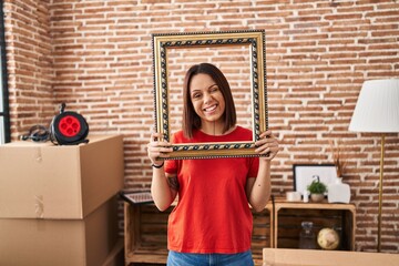 Young hispanic woman at new home holding empty frame winking looking at the camera with sexy...