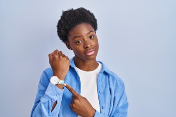 African american woman standing over blue background in hurry pointing to watch time, impatience,...