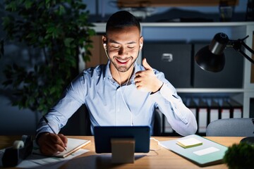 Young hispanic man working at the office at night doing happy thumbs up gesture with hand. approving expression looking at the camera showing success.