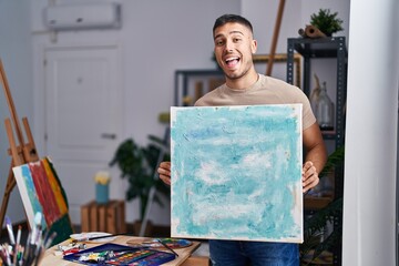 Young hispanic painter man holding canvas smiling and laughing hard out loud because funny crazy...