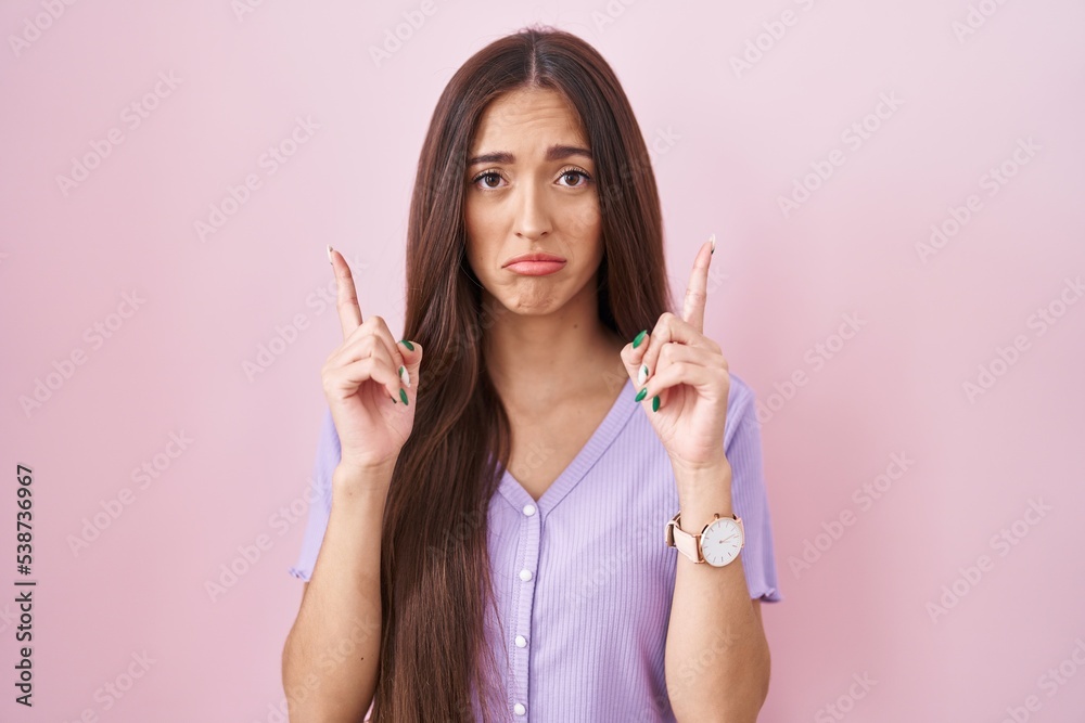 Sticker Young hispanic woman with long hair standing over pink background pointing up looking sad and upset, indicating direction with fingers, unhappy and depressed.