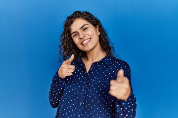 Young brunette woman with curly hair wearing casual clothes over blue background pointing fingers to camera with happy and funny face. good energy and vibes.