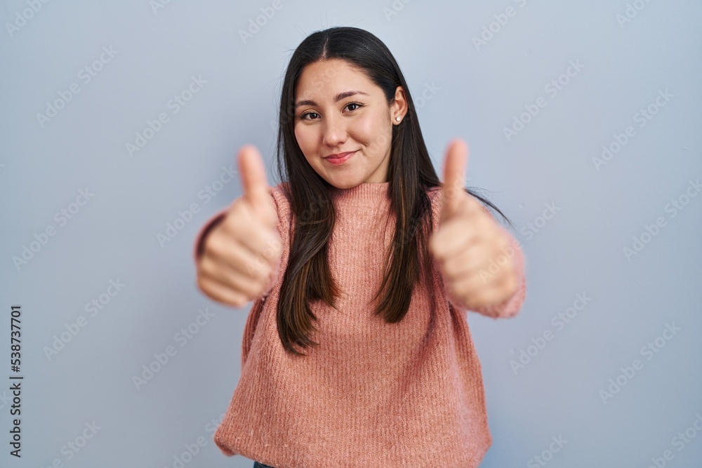 Wall mural Young latin woman standing over blue background approving doing positive gesture with hand, thumbs up smiling and happy for success. winner gesture.