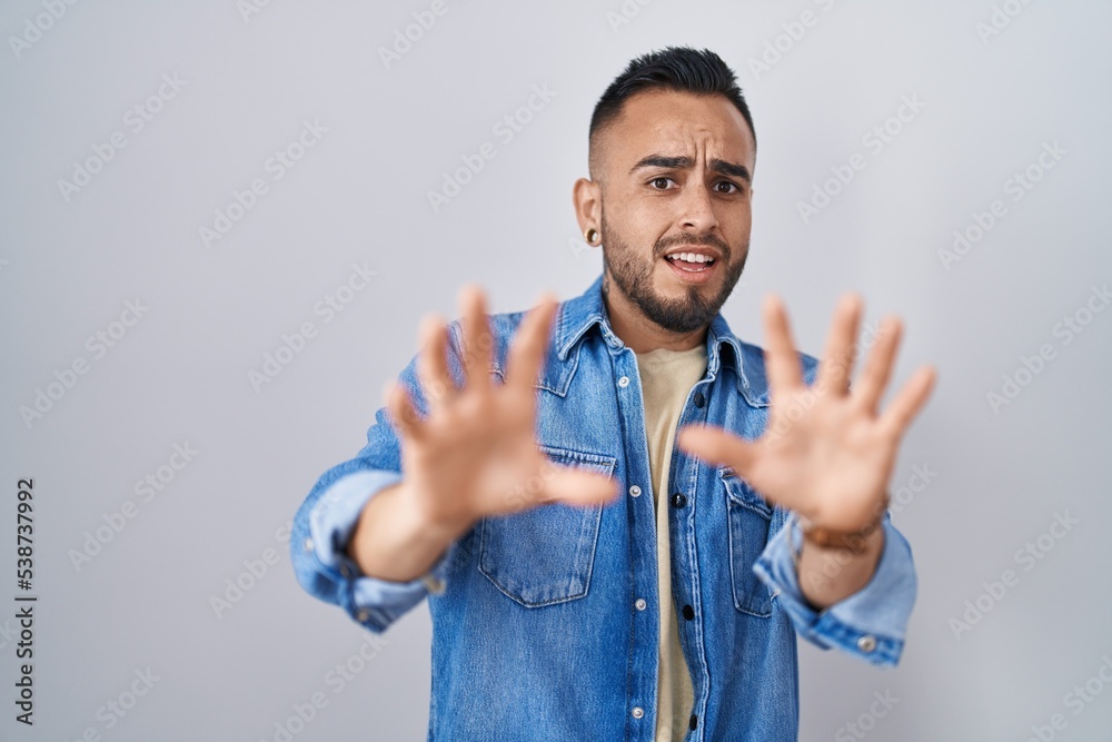 Sticker Young hispanic man standing over isolated background afraid and terrified with fear expression stop gesture with hands, shouting in shock. panic concept.