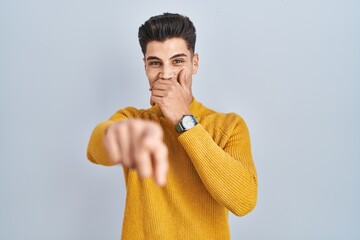 Young hispanic man standing over blue background laughing at you, pointing finger to the camera...