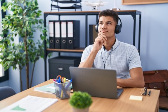 Young hispanic man working at the office wearing headphones with hand on chin thinking about question, pensive expression. smiling with thoughtful face. doubt concept.