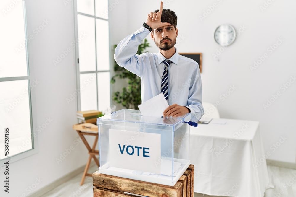 Sticker Hispanic man with beard voting putting envelop in ballot box making fun of people with fingers on forehead doing loser gesture mocking and insulting.