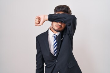 Young hispanic man with tattoos wearing business suit and tie covering eyes with arm, looking serious and sad. sightless, hiding and rejection concept