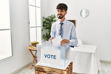 Hispanic man with beard voting putting envelop in ballot box winking looking at the camera with...