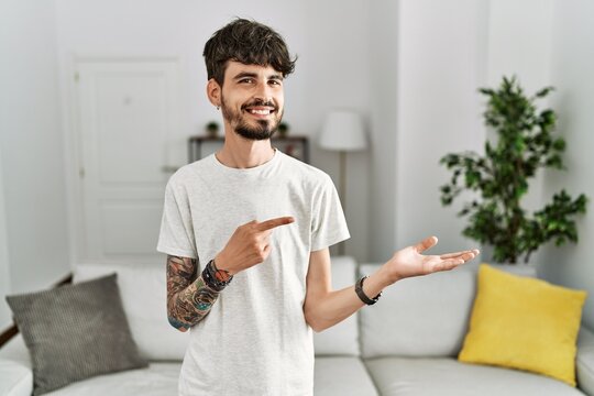 Hispanic man with beard at the living room at home amazed and smiling to the camera while presenting with hand and pointing with finger.