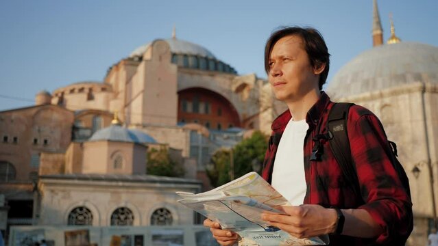 Man tourist walking near Ayasofya (Hagia Sophia) Mosque, looking at paper map, travel concept. Caucasian guy wearing shirt holding and navigating with topographic map, area in Istanbul Fatih