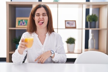 Brunette woman drinking glass of orange juice sticking tongue out happy with funny expression. emotion concept.