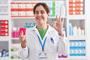 Brunette woman working at pharmacy drugstore holding condom doing ok sign with fingers, smiling...