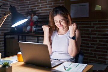 Brunette woman working at the office at night very happy and excited doing winner gesture with arms raised, smiling and screaming for success. celebration concept.