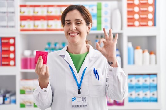 Brunette woman working at pharmacy drugstore holding condom doing ok sign with fingers, smiling friendly gesturing excellent symbol