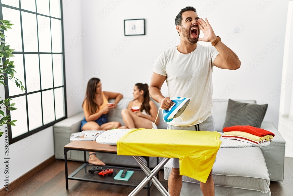 Poster young hispanic man ironing clothes at home shouting and screaming loud to side with hand on mouth. c