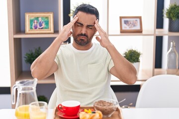 Hispanic man with beard eating breakfast with hand on head, headache because stress. suffering migraine.