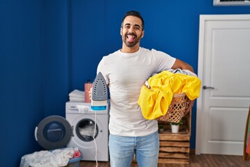 Young hispanic man with beard holding iron and clothes at home sticking tongue out happy with funny expression.