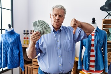 Senior man with grey hair holding dollars banknotes at retail shop with angry face, negative sign...
