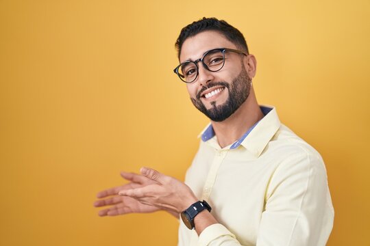 Hispanic young man wearing business clothes and glasses inviting to enter smiling natural with open hand