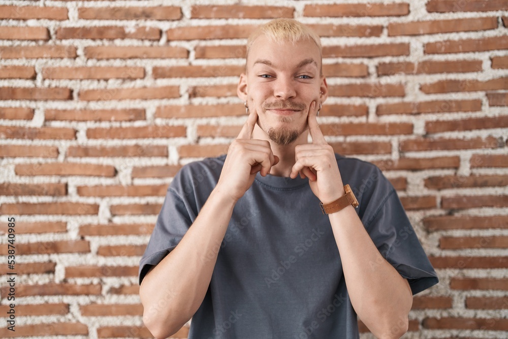 Poster young caucasian man standing over bricks wall smiling with open mouth, fingers pointing and forcing 