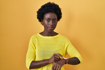 African young woman standing over yellow studio checking the time on wrist watch, relaxed and...