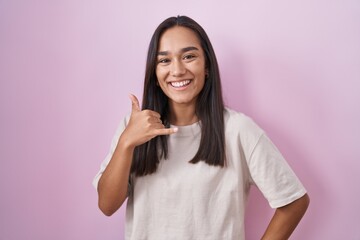 Young hispanic woman standing over pink background smiling doing phone gesture with hand and...
