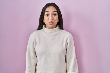 Young south asian woman standing over pink background puffing cheeks with funny face. mouth inflated with air, crazy expression.