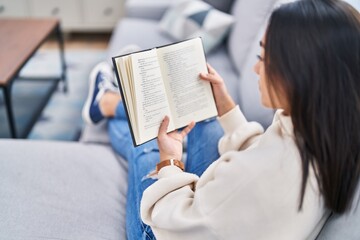 Young hispanic woman reading book sitting on sofa at home