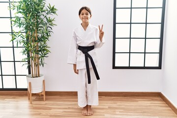 Young hispanic girl wearing karate kimono and black belt showing and pointing up with fingers number two while smiling confident and happy.