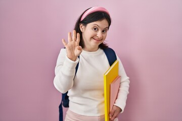 Woman with down syndrome wearing student backpack and holding books waiving saying hello happy and smiling, friendly welcome gesture
