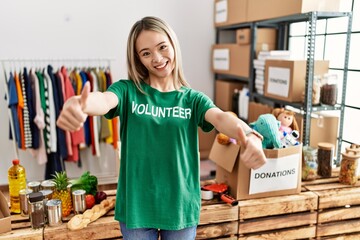 Asian young woman wearing volunteer t shirt at donations stand approving doing positive gesture with hand, thumbs up smiling and happy for success. winner gesture.