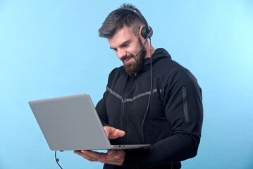Young bodybuilder, online coach, working on his laptop, consulting with a client through his headset; blue studio background