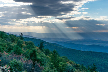 Rays of light can be seen shining through the clouds over the blue Ridge Mountains inside Shenandoah National Park. The foreground of the photo features the brush and forests of a mountain.